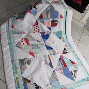 Könnte beinhalten: Eine weiße Steppdecke mit Patchwork-Muster in verschiedenen Farben und Mustern, darunter Rot, Blau, Grün und Grau. Die Steppdecke besteht aus Quadraten und Dreiecken und die Kanten sind mit einem hellblauen und weißen Band eingefasst.