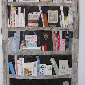Könnte beinhalten: Ein Wandbild aus Stoff, das ein Bücherregal mit verschiedenen bunten Stoffresten und Mustern darstellt. Das Bücherregal hat fünf Regalbretter und ist gefüllt mit Büchern, Stoffresten und anderen Dekorationsartikeln. Die Stoffreste sind in verschiedenen Mustern und Farben angeordnet, darunter Rot, Blau, Grün, Gelb und Weiß.