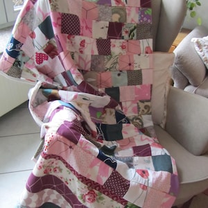 Könnte beinhalten: Eine handgefertigte Patchwork-Decke, die über einem Stuhl drapiert ist. Die Decke zeigt eine Vielzahl von Stoffen in Rosa-, Weiß- und Burgundertönen mit floralen und geometrischen Mustern. Die Decke ist eine Mischung aus Quadraten und Streifen und wirkt gemütlich.