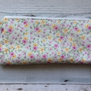 Puede incluir: Un estuche rectangular con estampado floral y cremallera blanca. El estuche presenta un fondo verde claro con un patr&oacute;n de flores rosas y amarillas. El tirador de la cremallera es plateado.