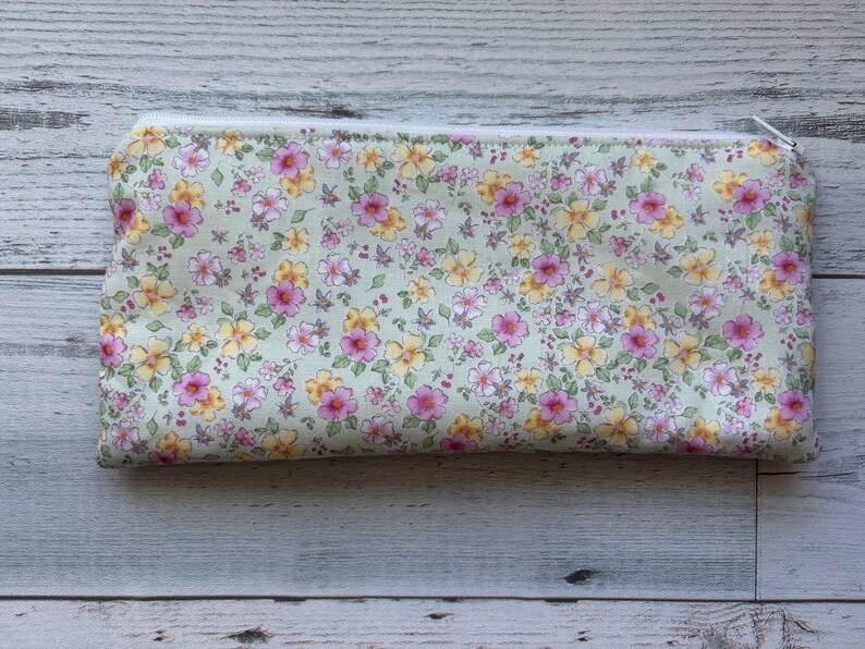 Puede incluir: Un estuche rectangular con estampado floral y cremallera blanca. El estuche presenta un fondo verde claro con un patr&oacute;n de flores rosas y amarillas. El tirador de la cremallera es plateado.