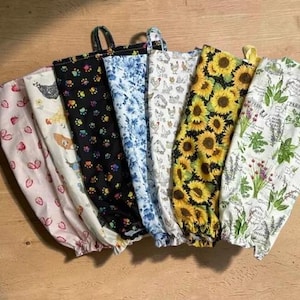 May include: A collection of six fabric bags with hanging loops, featuring various patterns including pink strawberries, chickens, colorful paw prints, blue floral designs, yellow sunflowers, and green herbs on white and black backgrounds.