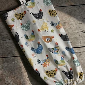 May include: A cream-colored fabric bag featuring a repeating pattern of stylized chickens in various colors including black, blue, yellow, and green. The bag has a gathered bottom opening and a fabric loop at the top for hanging storage.