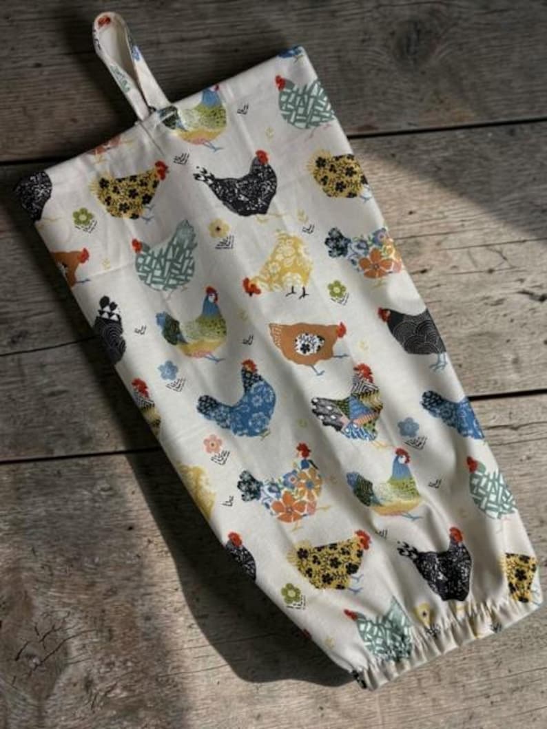 May include: A cream-colored fabric bag featuring a repeating pattern of stylized chickens in various colors including black, blue, yellow, and green. The bag has a gathered bottom opening and a fabric loop at the top for hanging storage.