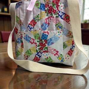 May include: A white tote bag with a colorful patchwork design featuring floral and geometric patterns. The bag has a cream-colored shoulder strap and a pink ribbon handle. The bag is sitting on a wooden table.