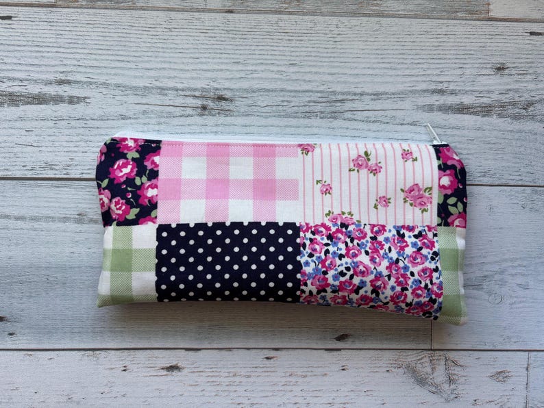 Puede incluir: Un estuche rectangular con un dise&ntilde;o de patchwork. El estuche presenta varios patrones de tela, incluyendo cuadros rosas, estampados florales, lunares y rayas. Una cremallera blanca recorre el borde superior. El estuche mide aproximadamente 20 cm de largo.