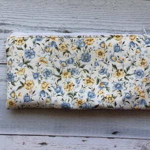 Puede incluir: Bolsa rectangular con estampado floral y cremallera blanca. La tela presenta un patr&oacute;n de flores azules y amarillas con hojas verdes sobre un fondo crema. La bolsa se utiliza probablemente para cosm&eacute;ticos o art&iacute;culos peque&ntilde;os.