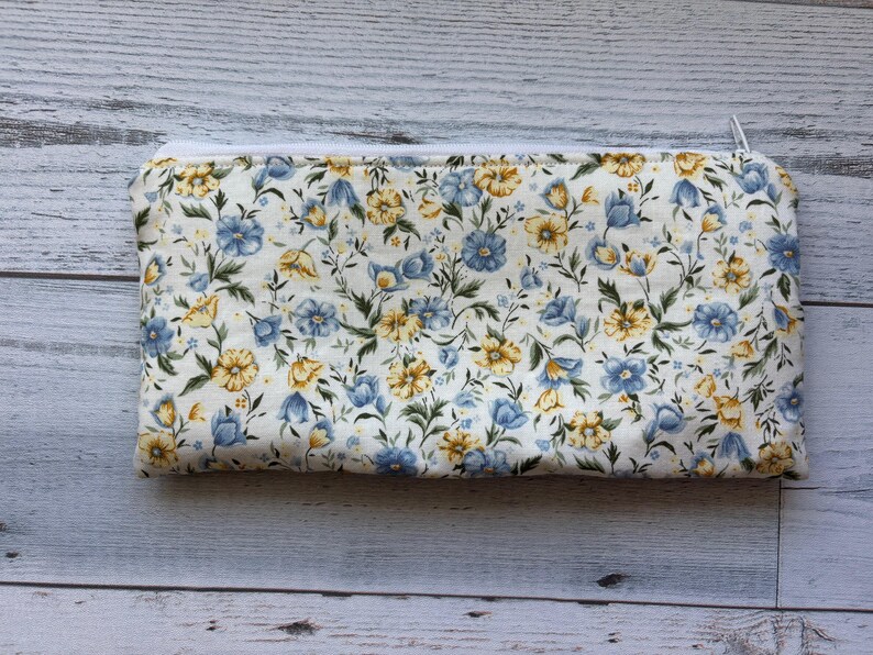 Puede incluir: Bolsa rectangular con estampado floral y cremallera blanca. La tela presenta un patr&oacute;n de flores azules y amarillas con hojas verdes sobre un fondo crema. La bolsa se utiliza probablemente para cosm&eacute;ticos o art&iacute;culos peque&ntilde;os.
