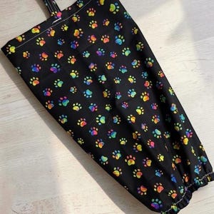 May include: A black fabric bag featuring a repeating pattern of colorful, rainbow-hued paw prints. The bag has a rectangular shape with a top hanging loop and a gathered, elasticized bottom opening for dispensing items through the base.