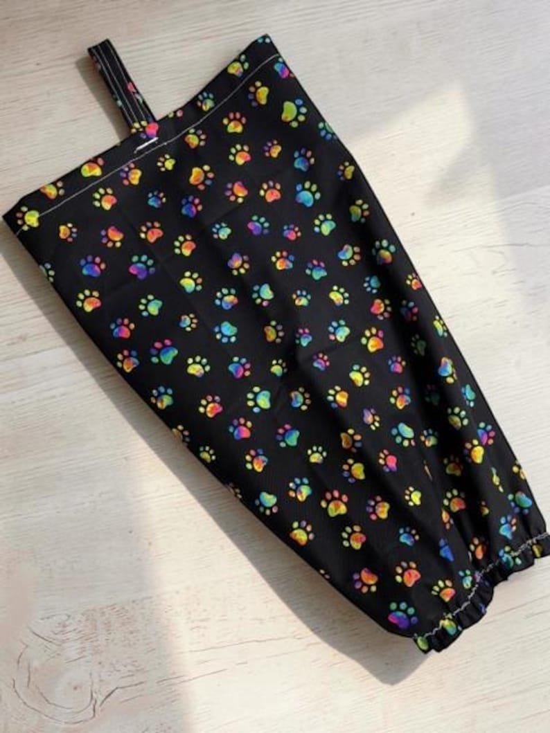 May include: A black fabric bag featuring a repeating pattern of colorful, rainbow-hued paw prints. The bag has a rectangular shape with a top hanging loop and a gathered, elasticized bottom opening for dispensing items through the base.