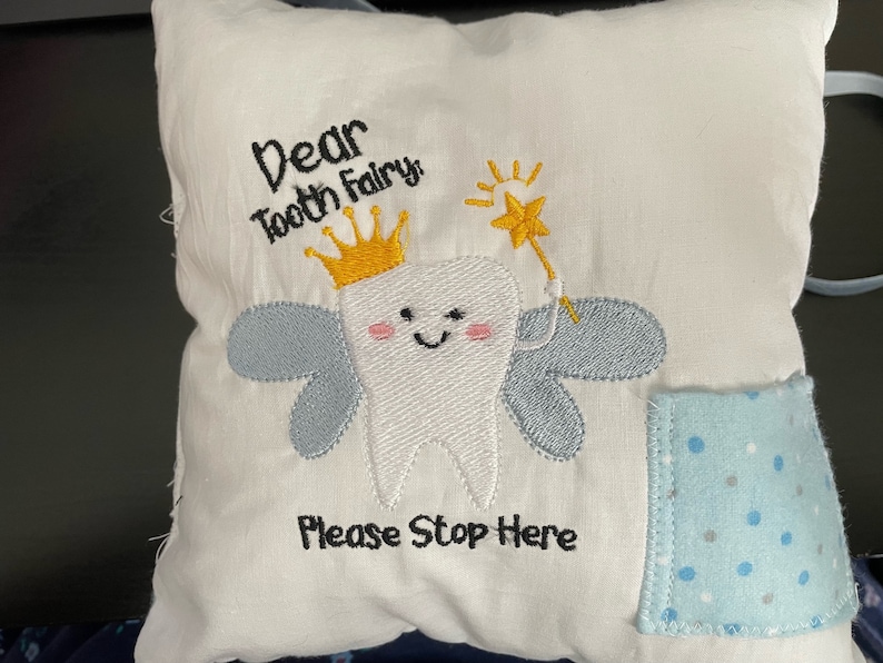 May include: White pillow with embroidered tooth fairy design. The tooth fairy is a white tooth with a yellow crown, wings, and a wand. The text reads "Dear Tooth Fairy Please Stop Here".