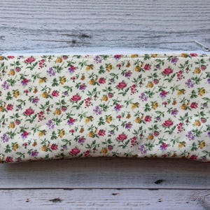 Puede incluir: Un estuche rectangular con estampado floral y cremallera blanca. El estuche presenta un fondo crema cubierto de peque&ntilde;as flores rosas, amarillas y moradas con hojas verdes. La cremallera es blanca.