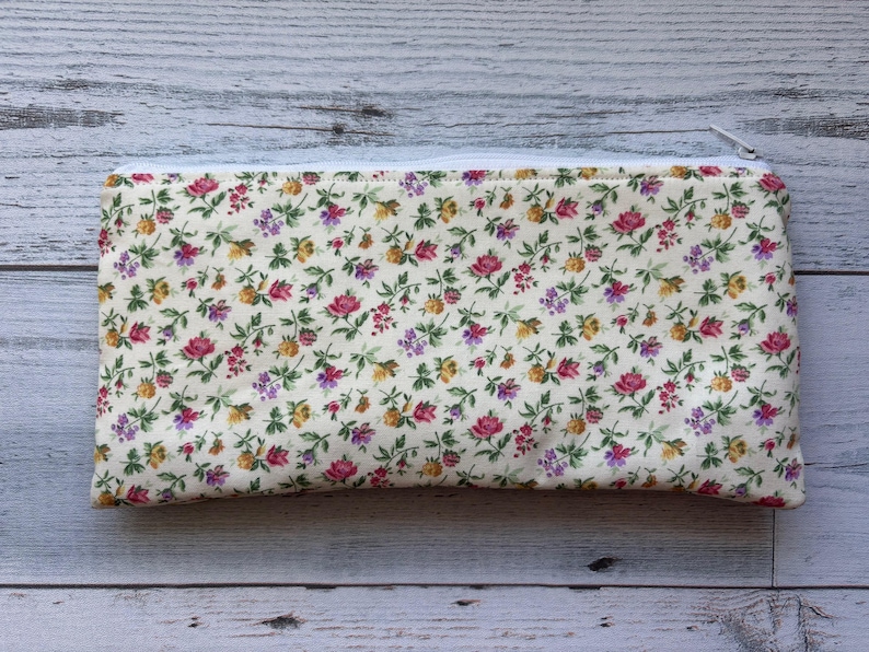 Puede incluir: Un estuche rectangular con estampado floral y cremallera blanca. El estuche presenta un fondo crema cubierto de peque&ntilde;as flores rosas, amarillas y moradas con hojas verdes. La cremallera es blanca.