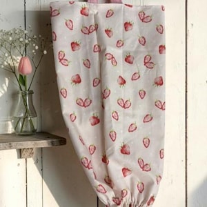 May include: A fabric bag featuring a repeating pattern of red strawberries on a light cream background with small white dots. The bag has a pink fabric handle at the top and an elasticized opening at the bottom for dispensing items.
