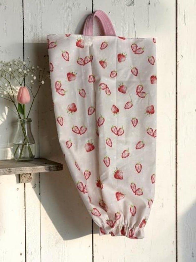 May include: A fabric bag featuring a repeating pattern of red strawberries on a light cream background with small white dots. The bag has a pink fabric handle at the top and an elasticized opening at the bottom for dispensing items.