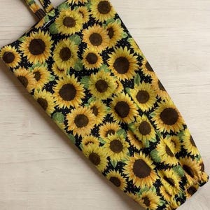 May include: A fabric bag featuring a repeating pattern of yellow sunflowers with brown centers on a black background. The bag has a rectangular shape, a fabric loop at the top for hanging, and an elasticized opening at the bottom edge.