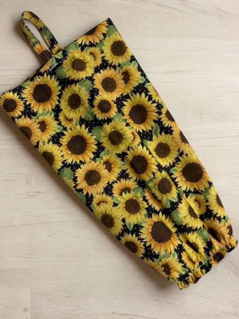 May include: A fabric bag featuring a repeating pattern of yellow sunflowers with brown centers on a black background. The bag has a rectangular shape, a fabric loop at the top for hanging, and an elasticized opening at the bottom edge.