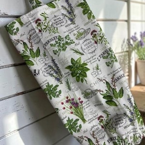 May include: A white fabric bag featuring a repeating botanical print of various green herbs including parsley, sage, and chives. The design includes black text labels such as Home Grown Herbs, Herb Garden, and Parsley, with a hanging loop at the top.