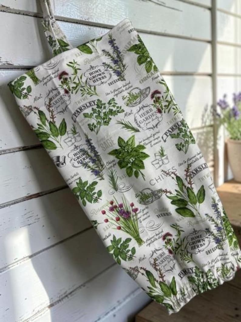 May include: A white fabric bag featuring a repeating botanical print of various green herbs including parsley, sage, and chives. The design includes black text labels such as Home Grown Herbs, Herb Garden, and Parsley, with a hanging loop at the top.