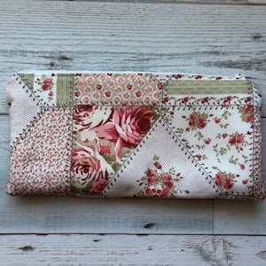 Puede incluir: Un estuche rectangular de tela patchwork con estampado floral. El dise&ntilde;o presenta varios estampados florales en tonos rosa, rojo y verde, sobre un fondo blanco. El estuche est&aacute; cosido con hilo en contraste, con un aspecto vintage.