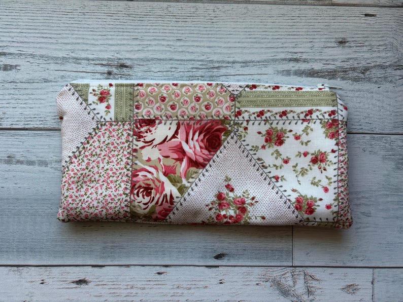 Puede incluir: Un estuche rectangular de tela patchwork con estampado floral. El dise&ntilde;o presenta varios estampados florales en tonos rosa, rojo y verde, sobre un fondo blanco. El estuche est&aacute; cosido con hilo en contraste, con un aspecto vintage.