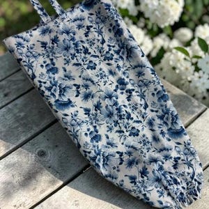 May include: A fabric bag featuring a blue and white floral pattern with a tapered shape, a top hanging loop, and an elasticized opening at the bottom. The textile displays a dense arrangement of various blossoms and leaves in shades of blue.