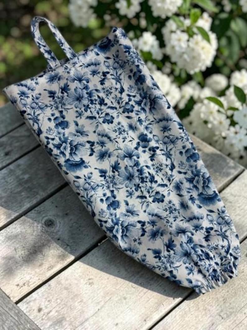 May include: A fabric bag featuring a blue and white floral pattern with a tapered shape, a top hanging loop, and an elasticized opening at the bottom. The textile displays a dense arrangement of various blossoms and leaves in shades of blue.
