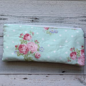 Puede incluir: Un estuche rectangular con un estampado floral. El estuche es de color azul claro con lunares blancos y presenta rosas rosas y flores moradas. Tiene una cremallera blanca.