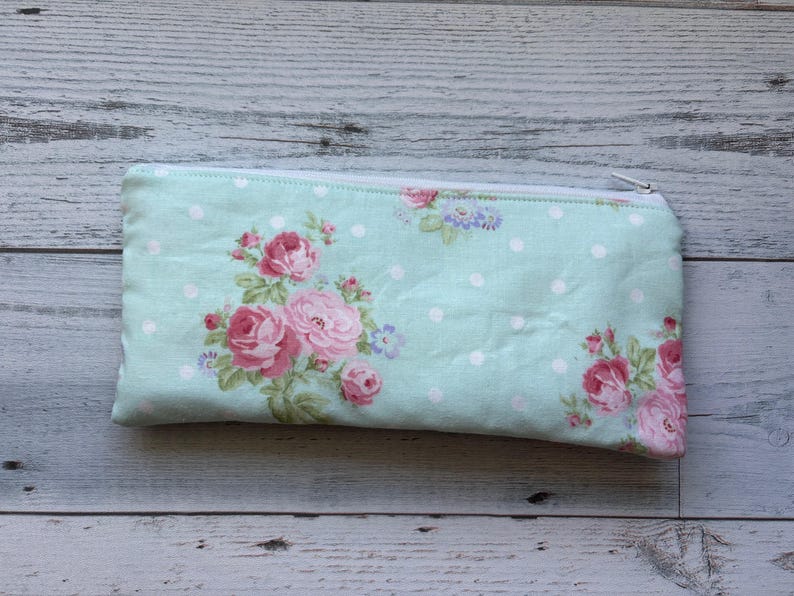 Puede incluir: Un estuche rectangular con un estampado floral. El estuche es de color azul claro con lunares blancos y presenta rosas rosas y flores moradas. Tiene una cremallera blanca.