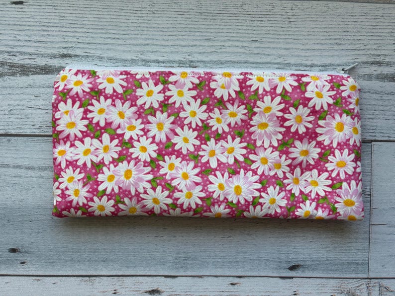 Puede incluir: Una bolsa rectangular con cremallera con un fondo rosa cubierto de margaritas blancas con centros amarillos. La bolsa est&aacute; hecha de tela y tiene una cremallera blanca.