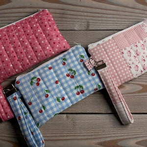 May include: Three quilted fabric pouches with wrist straps. One is pink with a heart pattern, another is blue gingham with cherry designs, and the third is pink with floral and gingham patches. Each has a zipper closure.
