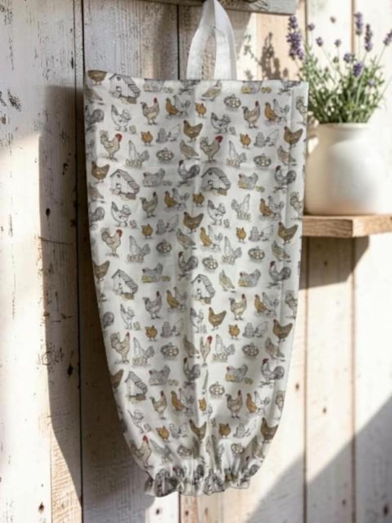 May include: A fabric bag with a white background featuring a repeating pattern of brown, grey, and white chickens, roosters, and small coops. The bag has a white fabric loop at the top for hanging and an elasticized opening at the bottom.