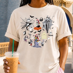 May include: A beige short-sleeved t-shirt featuring a graphic print of characters and motifs from The Nightmare Before Christmas, including Jack Skellington, Sally, Oogie Boogie, Zero, spiderwebs, bats, and pumpkins arranged in a central cluster.