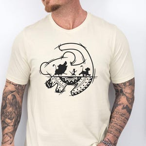 May include: Cream-colored t-shirt featuring a black graphic of a lion's head with three characters inside. The design includes a stylized outline and decorative patterns. The shirt is short-sleeved and has a crew neck.