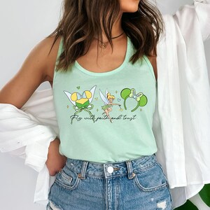 May include: A light green tank top featuring a graphic of a fairy, two sets of mouse ears, and the text "Fly with faith and trust" in a cursive font. The tank top is worn with a white button-down shirt and blue denim shorts with distressed details.