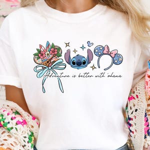 May include: White t-shirt featuring a colorful graphic of Stitch, a ukulele, and Mickey Mouse ears. The design includes the text "Adventure is better with 'ohana" in a script font. The shirt is a casual, everyday wear item.