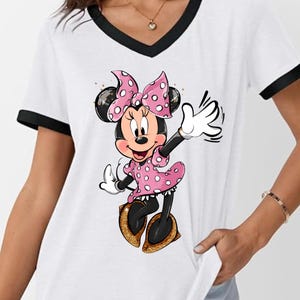 K&ouml;nnte beinhalten: Wei&szlig;es V-Ausschnitt-T-Shirt mit schwarzem Besatz mit einem Cartoon-Minnie-Mouse-Design. Minnie Mouse tr&auml;gt ein rosa gepunktetes Kleid und eine Schleife mit goldenen Glitzerschuhen. Das Shirt ist ein l&auml;ssiger Stil.