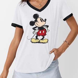 May include: White V-neck t-shirt with black trim featuring a graphic of Mickey Mouse. The cartoon character is depicted in his classic outfit: red shorts, yellow shoes, and white gloves. The shirt has short sleeves.