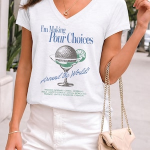 May include: A white V-neck t-shirt featuring a graphic of a margarita glass with a lime wedge and the text "I'm Making Pour Choices Around the World!" followed by a list of countries including Mexico, Norway, China, Germany, Italy, United States, Japan, Morocco, France, United Kingdom, and Canada.
