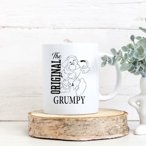 Pode incluir: Caneca de cerâmica branca com uma ilustração em preto e branco do Grumpy da Branca de Neve. O texto "The Original Grumpy" está impresso na caneca.