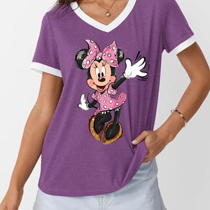 K&ouml;nnte beinhalten: Lilafarbenes V-Ausschnitt-T-Shirt mit wei&szlig;em Besatz und Minnie Mouse-Grafik. Minnie tr&auml;gt ein rosa gepunktetes Kleid, eine Schleife und goldene Schuhe. Die Zeichentrickfigur winkt mit einer wei&szlig;en behandschuhten Hand.