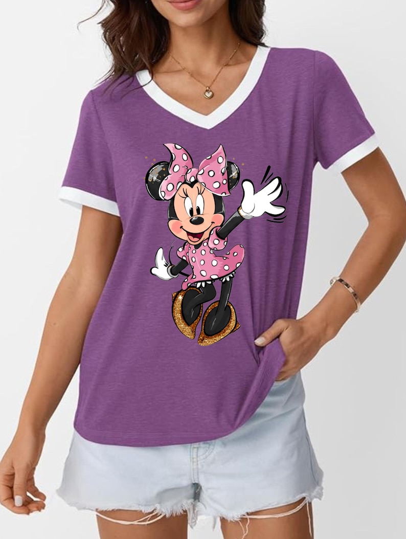 K&ouml;nnte beinhalten: Lilafarbenes V-Ausschnitt-T-Shirt mit wei&szlig;em Besatz und Minnie Mouse-Grafik. Minnie tr&auml;gt ein rosa gepunktetes Kleid, eine Schleife und goldene Schuhe. Die Zeichentrickfigur winkt mit einer wei&szlig;en behandschuhten Hand.