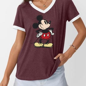 May include: Maroon V-neck t-shirt with white trim featuring a full-body graphic of Mickey Mouse. Mickey is wearing red shorts, yellow shoes, and white gloves. The shirt is short-sleeved and has a relaxed fit.