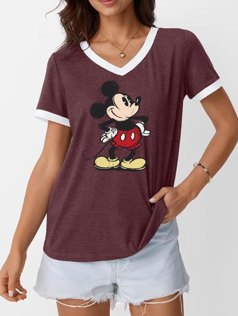 May include: Maroon V-neck t-shirt with white trim featuring a full-body graphic of Mickey Mouse. Mickey is wearing red shorts, yellow shoes, and white gloves. The shirt is short-sleeved and has a relaxed fit.