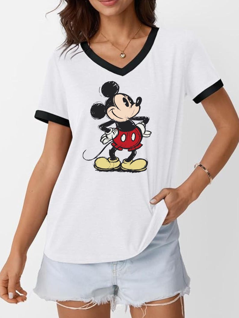 May include: White V-neck t-shirt with black trim featuring a graphic of Mickey Mouse. The cartoon character is depicted in his classic outfit: red shorts, yellow shoes, and white gloves. The shirt has short sleeves.