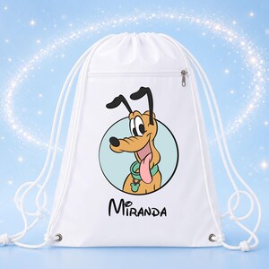 May include: A white drawstring backpack featuring a graphic of a cartoon dog with long black ears, a green collar, and a pink tongue inside a light blue circle. Below the graphic, the name Miranda is printed in black, stylized, cursive lettering.