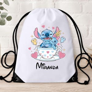 Op de afbeelding: Een witte rugzak met trekkoord voorzien van een afbeelding van het blauwe personage Stitch die een pop knuffelt in een theekopje versierd met hartjes. De naam Miranda staat in zwarte letters onder het kopje, omringd door roze hartjes en gele sterren.