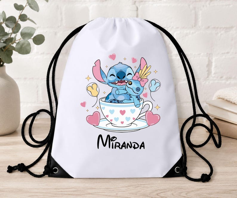 Op de afbeelding: Een witte rugzak met trekkoord voorzien van een afbeelding van het blauwe personage Stitch die een pop knuffelt in een theekopje versierd met hartjes. De naam Miranda staat in zwarte letters onder het kopje, omringd door roze hartjes en gele sterren.