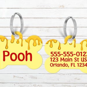 May include: Two yellow bone-shaped pet tags with silver key rings. One tag has the name "Pooh" in red. The other tag has a phone number and address in red. Both tags have a honey-like design.