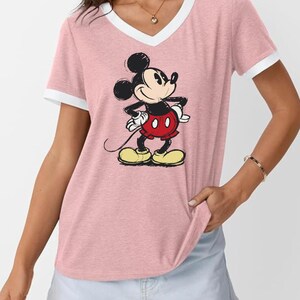 May include: Pink V-neck t-shirt with white trim featuring a cartoon illustration of Mickey Mouse. Mickey is depicted in his classic outfit: red shorts, yellow shoes, and white gloves. The shirt has short sleeves.